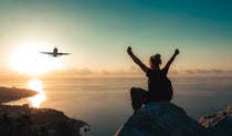 Woman sitting on top of a mountain with arms outstretched watching an aircraft fly overhead