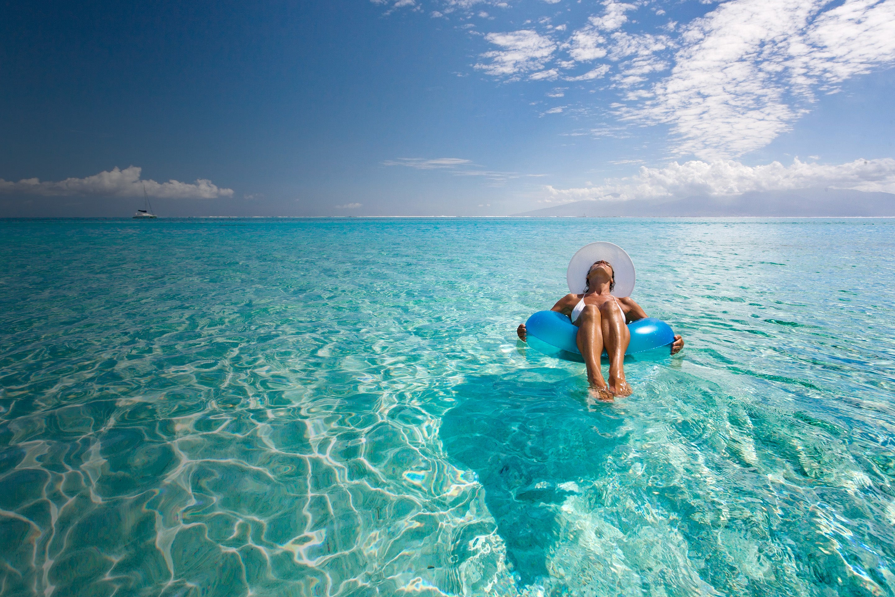 Woman sitting in an inflatable ring in the shallow waters of the sea with a yacht in the distance