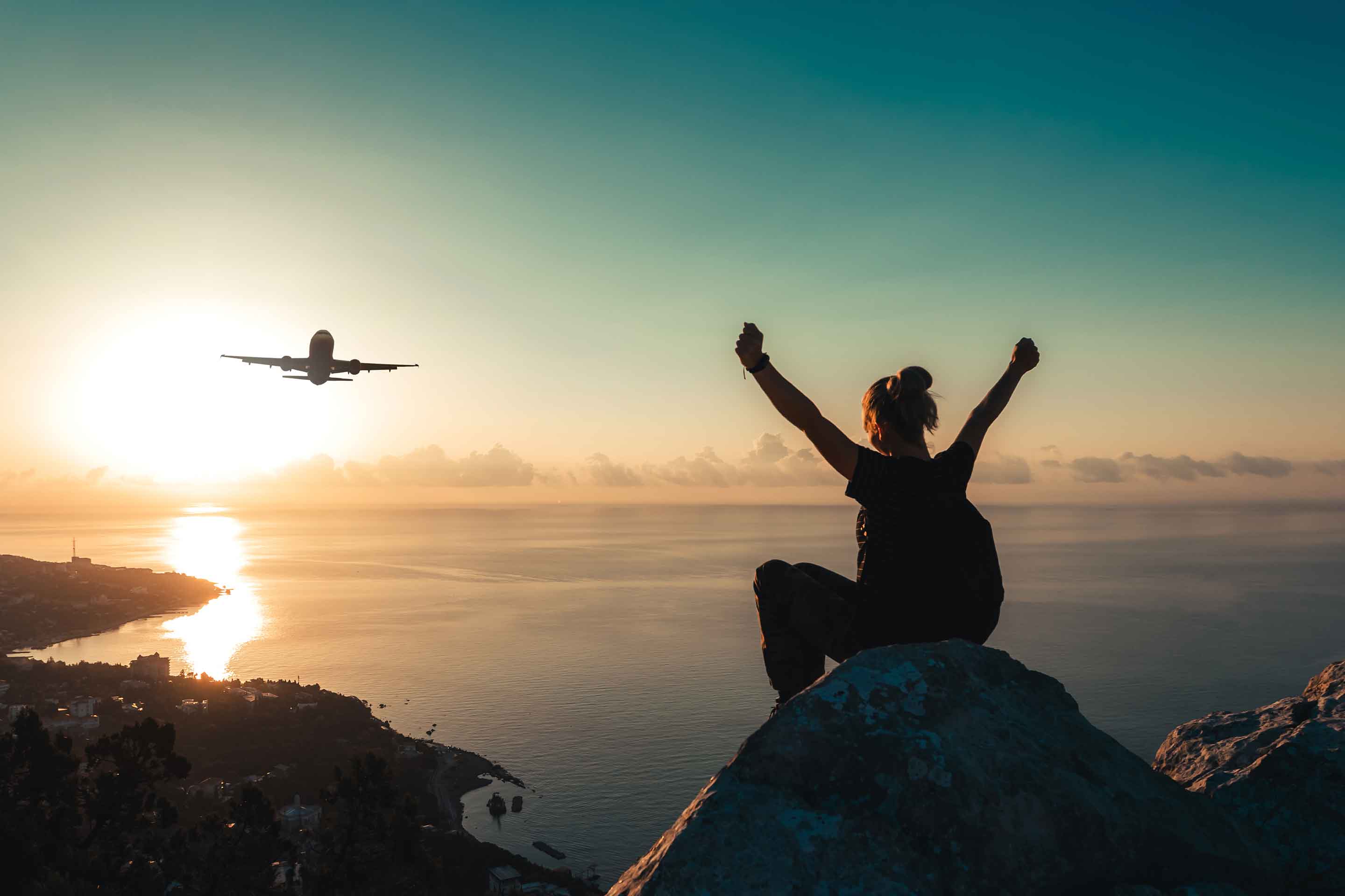 Woman sitting on top of a mountain with arms outstretched watching an aircraft fly overhead